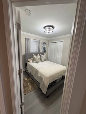 A bedroom with a bed, a nightstand, a window with curtains, a ceiling light fixture, and a door.