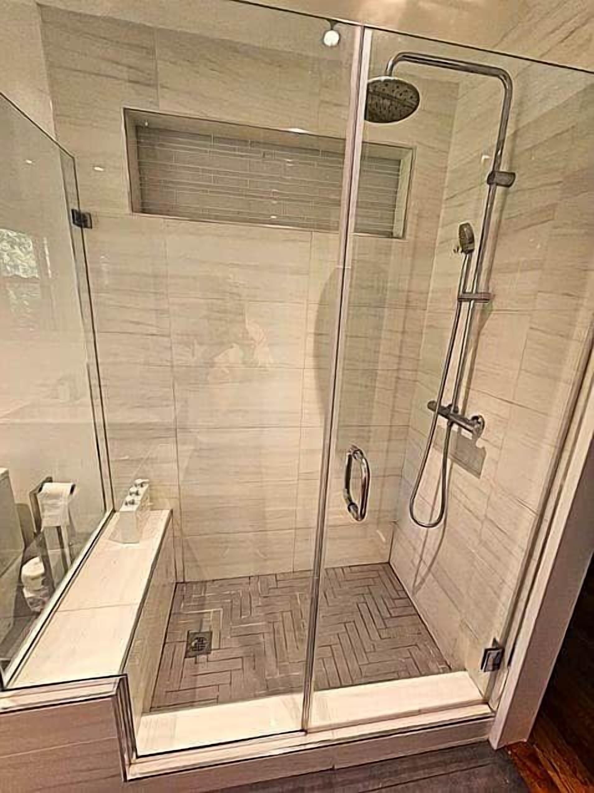 Un box de ducha con paredes de vidrio, un piso con estampado de espiga y un cabezal de ducha de mano, junto con un panel de control en la pared.