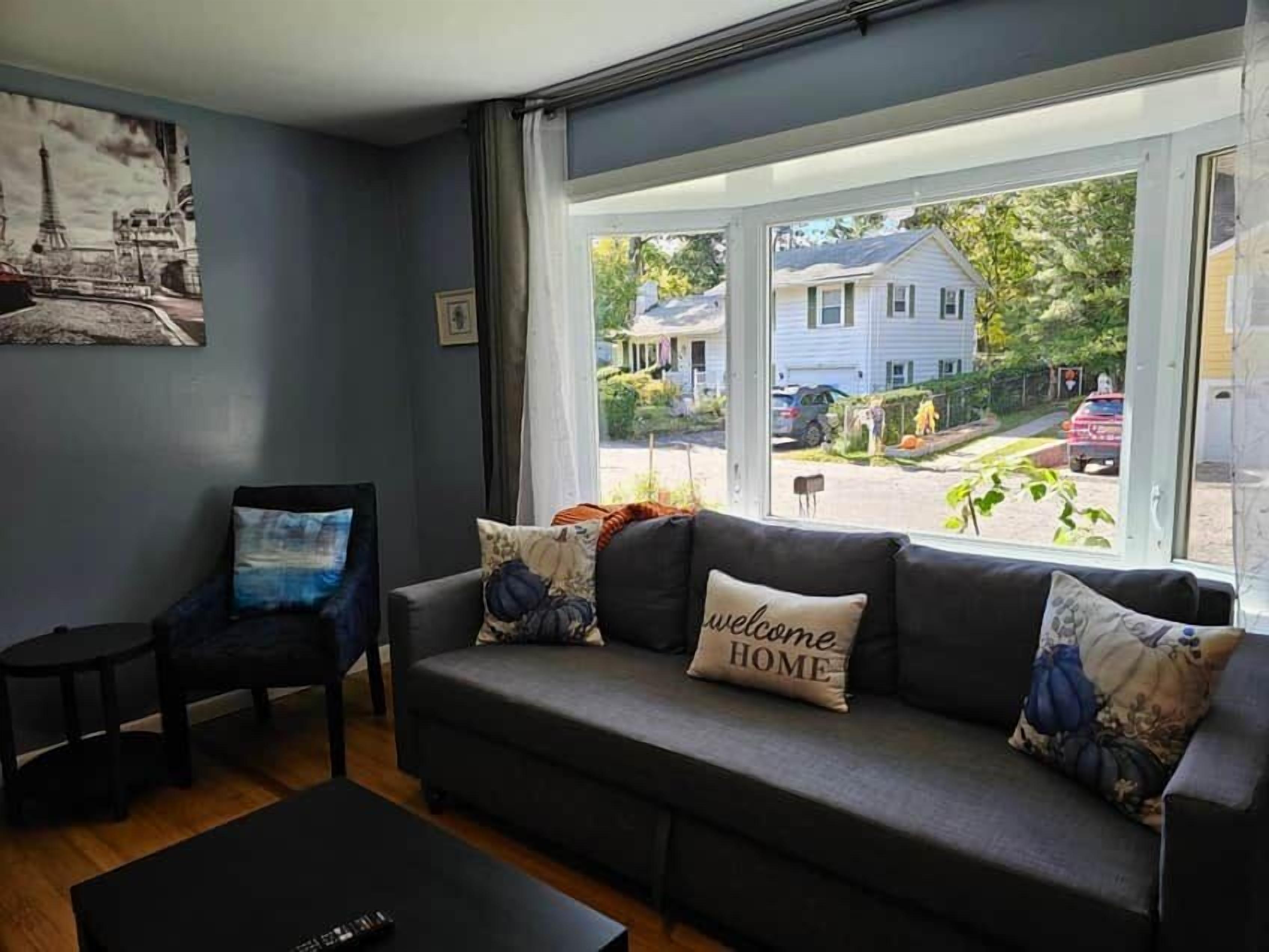 A living room with a sofa, a chair, and a coffee table. There is a window with a view of houses and trees.