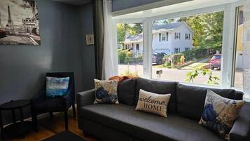 A living room with a sofa, a chair, and a coffee table. There is a window with a view of houses and trees.