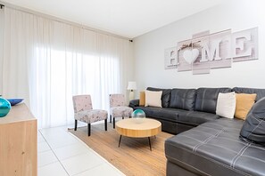 A modern living room with a black leather sofa, two armchairs, a wooden coffee table, and a large 'HOME' sign on the wall. - Disney 4BR Home + Pool & Themed Bedrooms (Kissimmee)