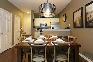 A dining area with a wooden table set for four, a kitchen in the background, and a large pendant light above. - 3BR West Lucaya Home With Clubhouse (Kissimmee)