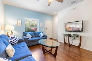 A living room with a blue sofa, a glass coffee table, a television, and a window with blinds. - 3BR Home With Resort Pool & Game Room (Kissimmee)