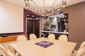 Un espace repas avec une grande table, des chaises, un lustre et une cuisine en arrière-plan.