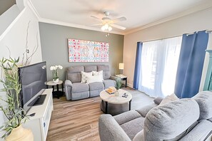 A modern living room with a grey sofa, a coffee table, and a TV. There is a ceiling fan, a painting on the wall, and a window with curtains. - 4BR Themed Townhome in Lucaya Resort (Kissimmee)