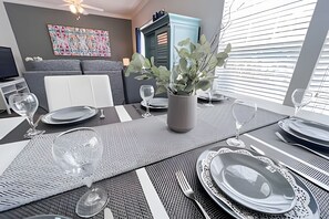 Una mesa de comedor dispuesta con platos, vasos y cubiertos, un florero con plantas verdes, y un sofá moderno al fondo.