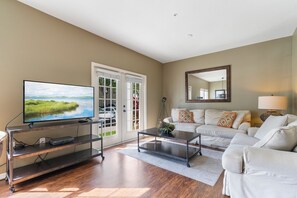A living room with a flat-screen TV on a metal stand, a white sofa, a coffee table, and a large mirror on the wall. - 4BR Lucaya Village Home, Sleeps 10 (Kissimmee)