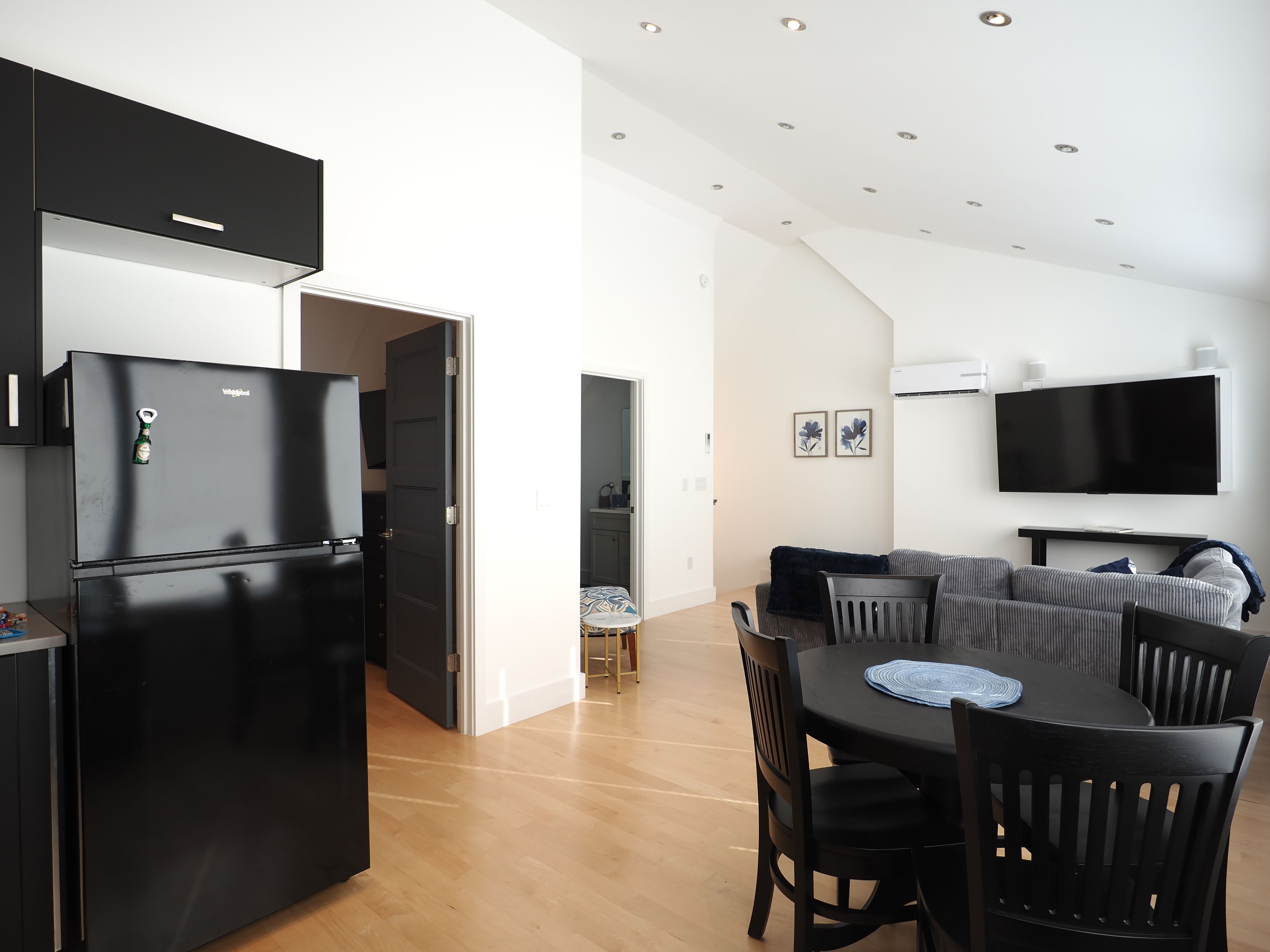 Eine moderne Küche mit einem schwarzen Kühlschrank, ein Essbereich mit einem runden Tisch und Stühlen sowie eine Wohnbereits mit einer Couch und einem Flachbildfernseher.