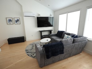 A modern living room with a gray sofa, a flat-screen TV, and a large window. - Downtown Cary Park View Bungalow (Cary)
