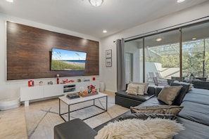 Een moderne woonkamer met een grote flatscreen televisie aan een houten wand, een bank met decoratieve kussens, een salontafel en een uitzicht door grote schuifdeuren op een terras.