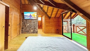 A wooden cabin room with a bed, a stone wall, a television, and a view of a green landscape.