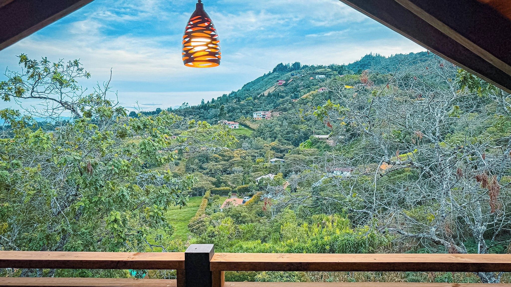 Vista desde un balcón con una lámpara colgante, baranda de madera y un paisaje de colinas con edificios dispersos.