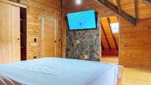 A bedroom with a bed, wooden walls, a stone wall, a television, and an open door leading to another room.