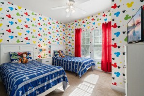 Ein Zimmer mit zwei Betten, einem Deckenventilator, einem Fenster mit roten Vorhängen und Wandstickern mit Mickey Mouse Motiven.