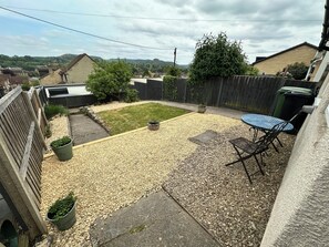 A backyard with a gravel patio, a small lawn, a wooden fence, and a table with chairs. - Cosy Cottage on the Cotswold way with Amazing views.  (Dursley)