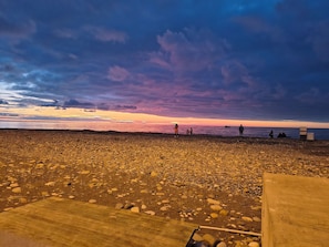 A beach with a wooden walkway leading to the water, people sitting and standing on the shore, and a sunset in the background. - Vrbo Property (Batumi)