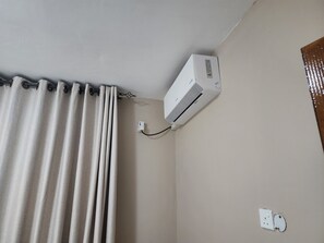 A room with a white air conditioner mounted on the wall, a curtain, and a power outlet.