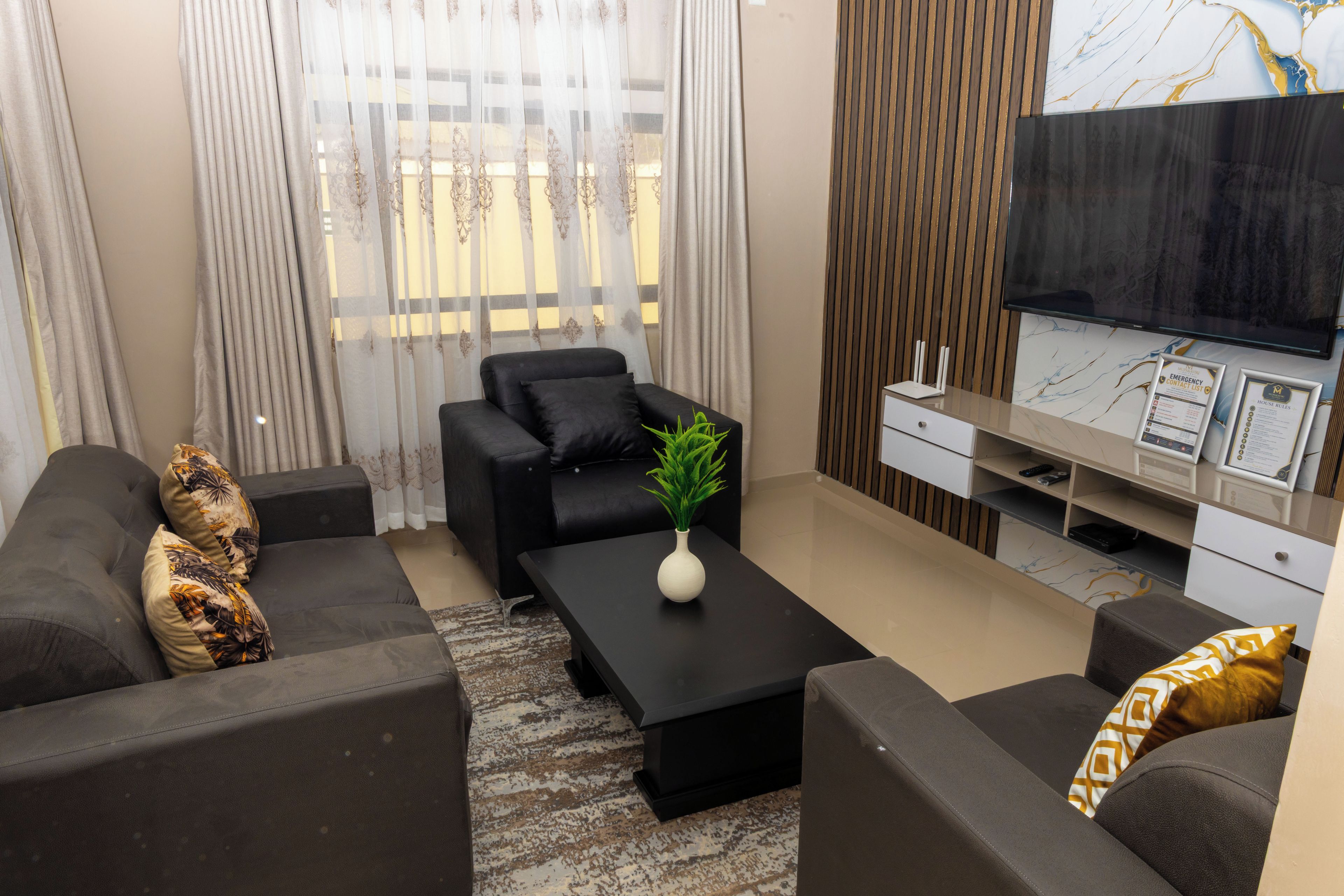 A modern living room with a black sofa set, a coffee table with a plant, a flat-screen TV, and a wooden accent wall.