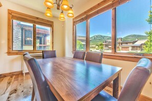 A dining area with a large wooden table and chairs, large windows with a view of buildings and hills, and a chandelier. - Luxury Pet Friendly 4-bed PC Ski & Lake House (Wasatch County)