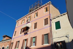A multi-story building with a pink facade, multiple windows with shutters, and a visible balcony. - Biennale Pano-view (Venice)