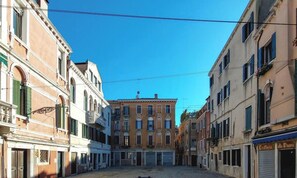 A narrow street with buildings on either side, a clear blue sky, and a central building with a prominent entrance. - Biennale Pano-view (Venice)