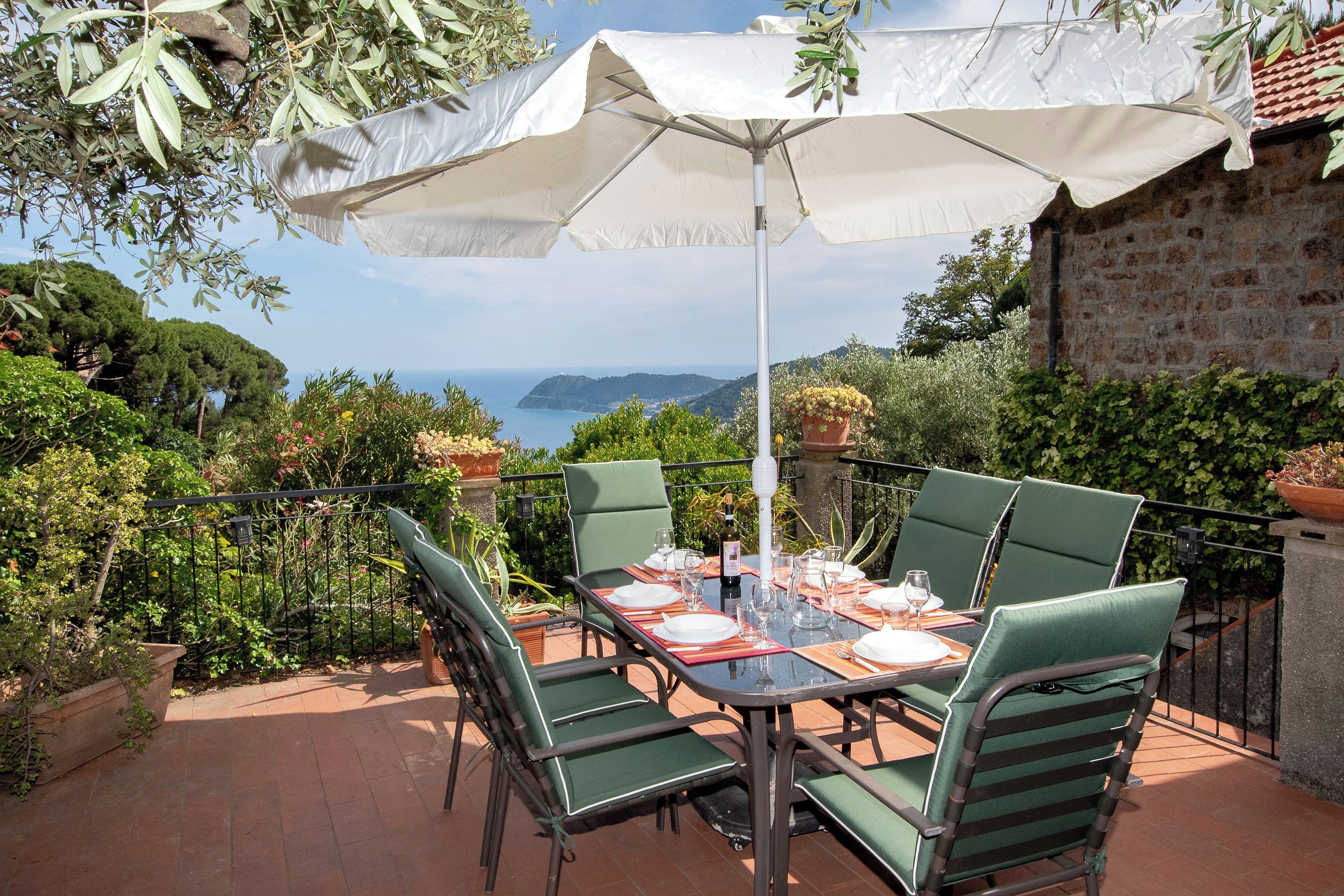 An outdoor dining area with a table set for four, surrounded by green chairs, under a white umbrella.