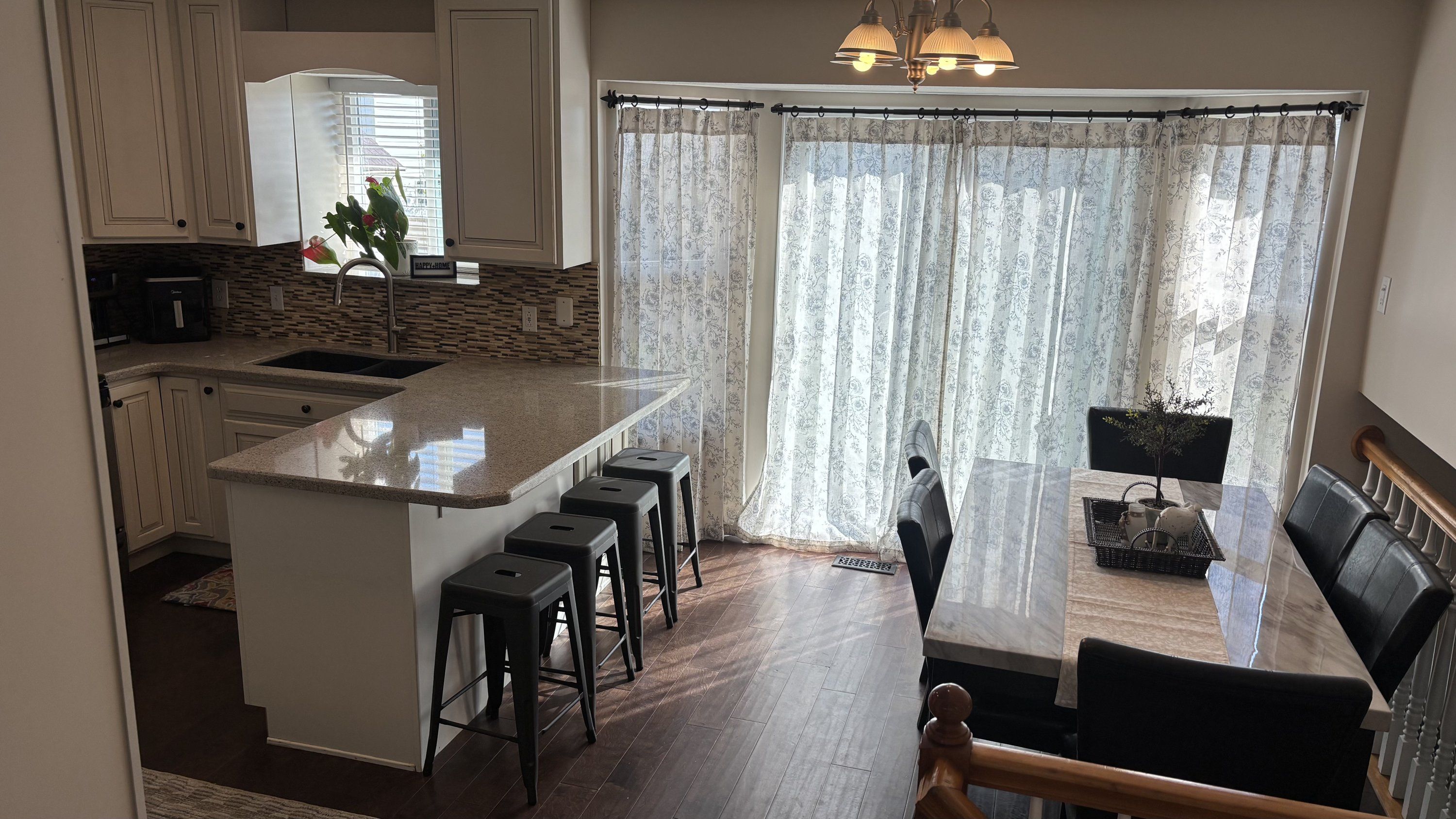 A modern kitchen with a central island, bar stools, a dining area with a table and chairs, and a view through a window with floral curtains.