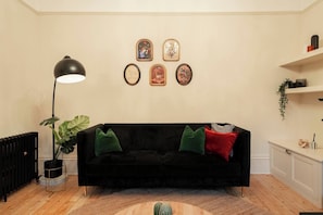 A living room with a black sofa, green and red cushions, a floor lamp, and wall-mounted picture frames. - The Gathering House - Beautiful Large House Suits Contractors (Leeds)