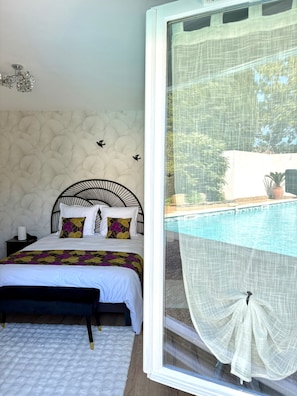 A bedroom with a bed, a nightstand, a decorative wall, and a window overlooking a pool. - La Closerie (La Redorte)
