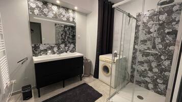 A bathroom with a shower, a washing machine, a sink, and a mirror.
