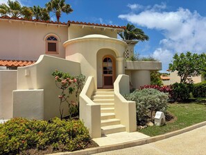 A Mediterranean-style building with a stucco exterior, a central arched entrance, and a small staircase leading to the door. - Palm Breeze Villa – Private Pool, Beach Club Access & Sugar Hill Resort (Porters)