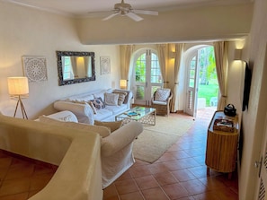 A spacious living room with a sofa, coffee table, and a television. There are two doors leading to outdoor areas. - Palm Breeze Villa – Private Pool, Beach Club Access & Sugar Hill Resort (Porters)