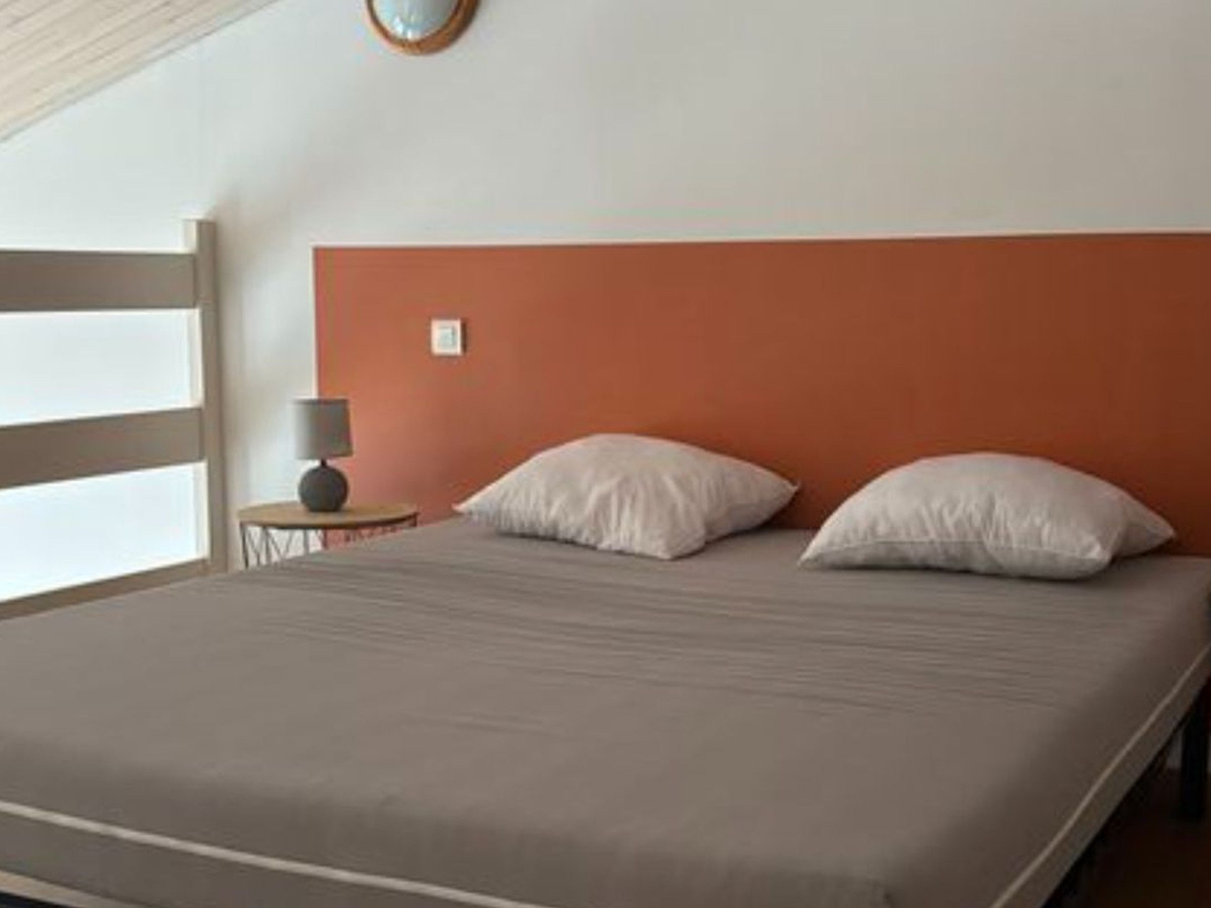 A bedroom with a bed, pillows, a nightstand, a lamp, and a wall clock.