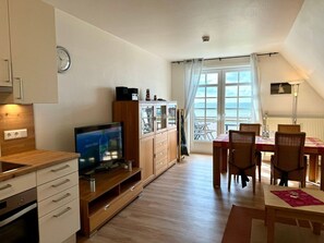 A compact living space with a kitchenette, a dining area with a table and chairs, a television, and a view of the sea through a large window. - "haus Parkstrasse" - Apt. 07 (Wyk auf Foehr)