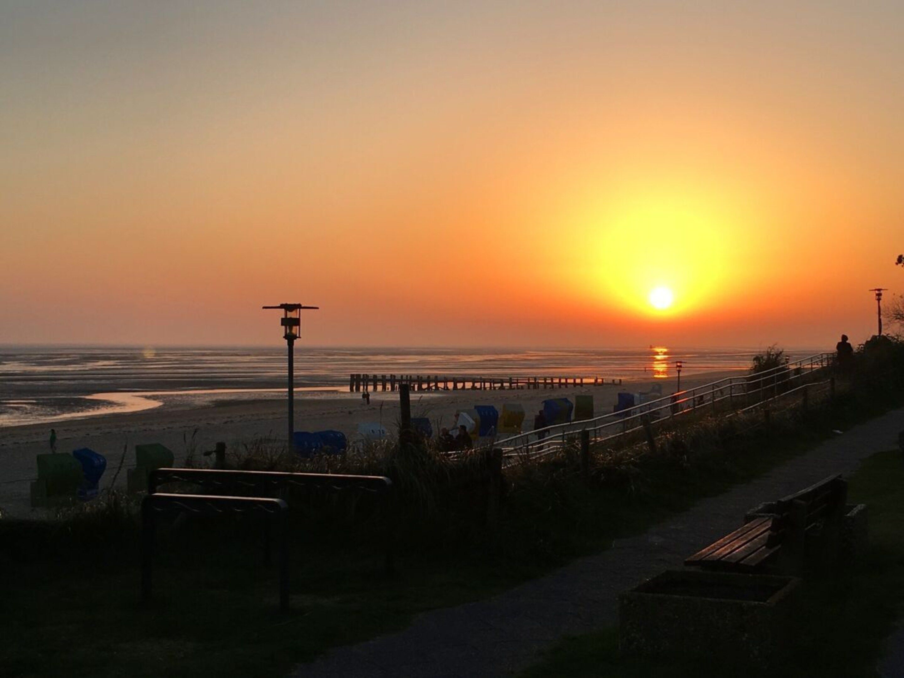 Ein Sonnenuntergang über einem Strand mit einem Steg, Bänken und einem Gehweg.