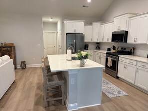 A modern kitchen with white cabinetry, a central island, stainless steel appliances, and a wicker chair. - Wonderful 4-bedroom house in adorable Pawleys Island (Pawleys Island)