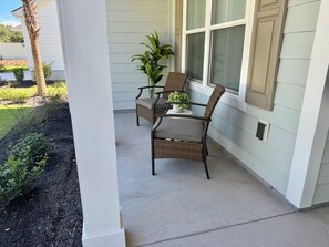 Eine überdachte Veranda mit zwei Korbstühlen, einem kleinen Tisch, einem Blumentopf und einem Fenster mit Fensterläden.