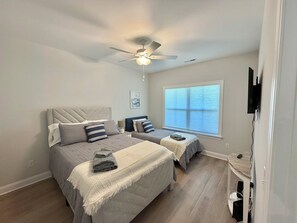 Ein Schlafzimmer mit zwei Betten, einem Deckenventilator, einem Fenster mit Jalousien und einem an der Wand befestigten Fernseher.