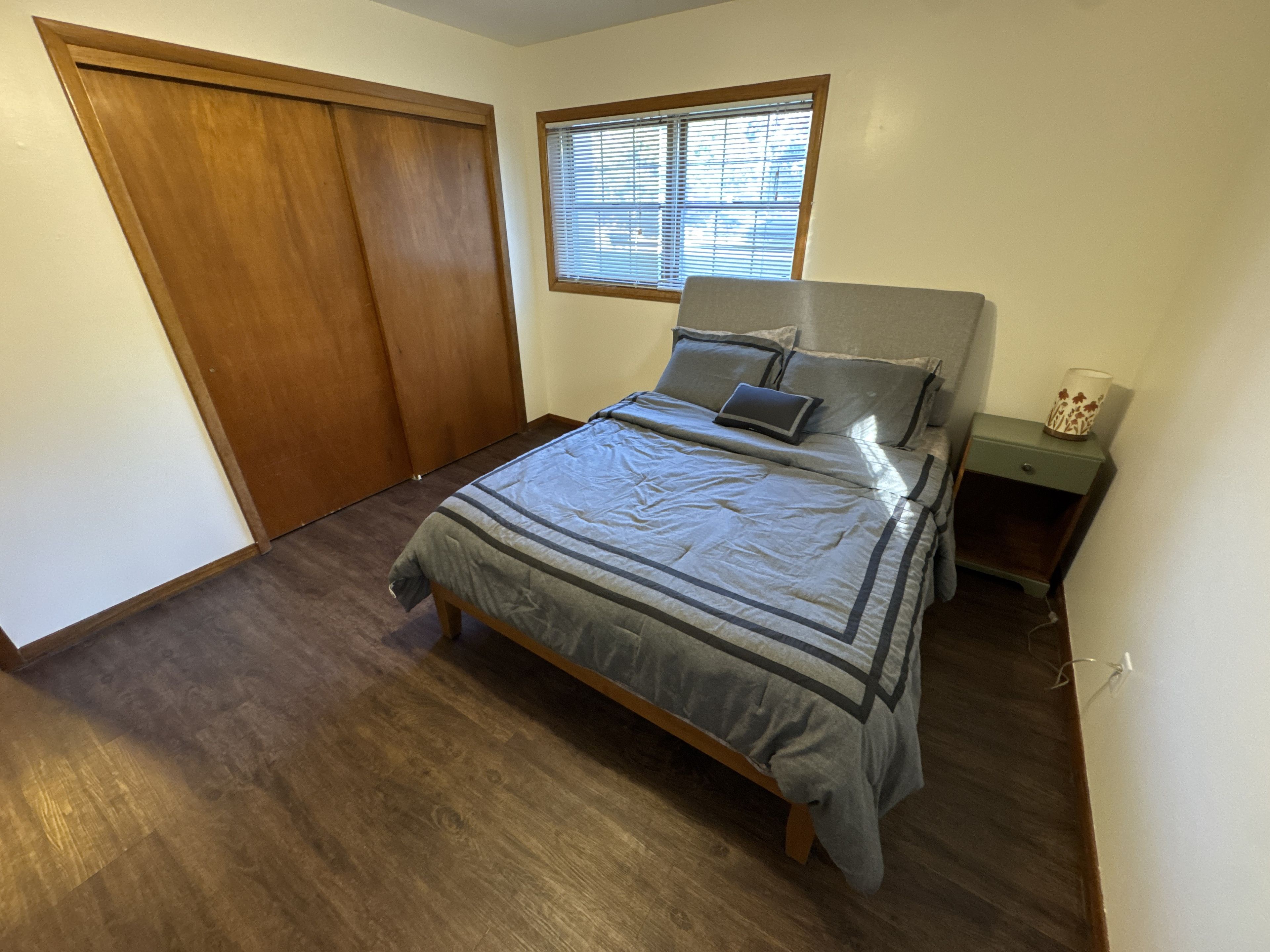 A bedroom with a bed, a nightstand, a window with blinds, and a wooden wardrobe.