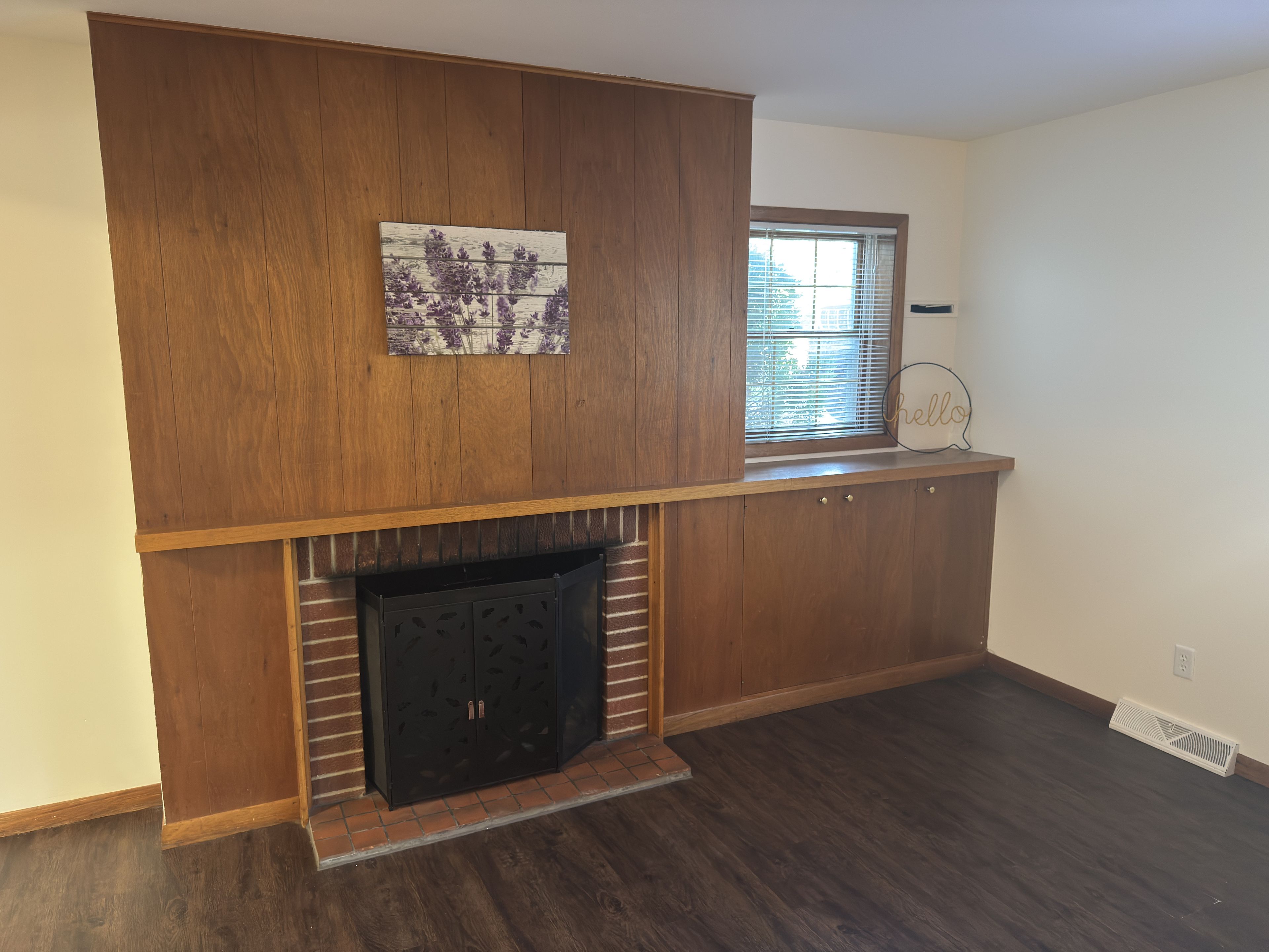 A room with a built-in fireplace, wooden paneling, a window with blinds, and a wall-mounted picture.