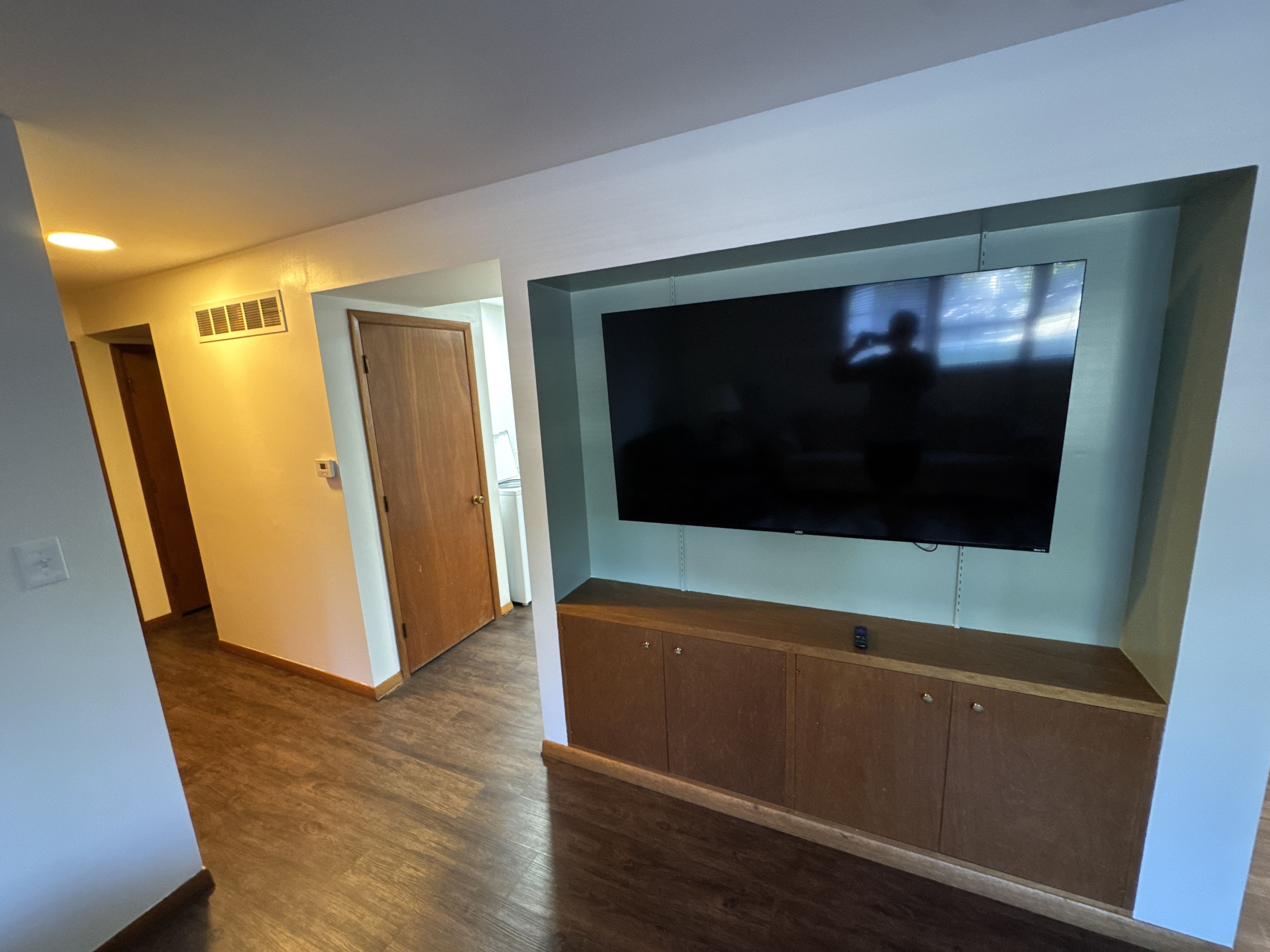 A living room with a flat-screen TV mounted on a wooden cabinet, a door leading to another room, and a hallway with wooden flooring.