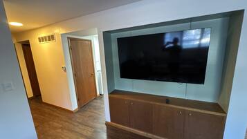 A living room with a flat-screen TV mounted on a wooden cabinet, a door leading to another room, and a hallway with wooden flooring.