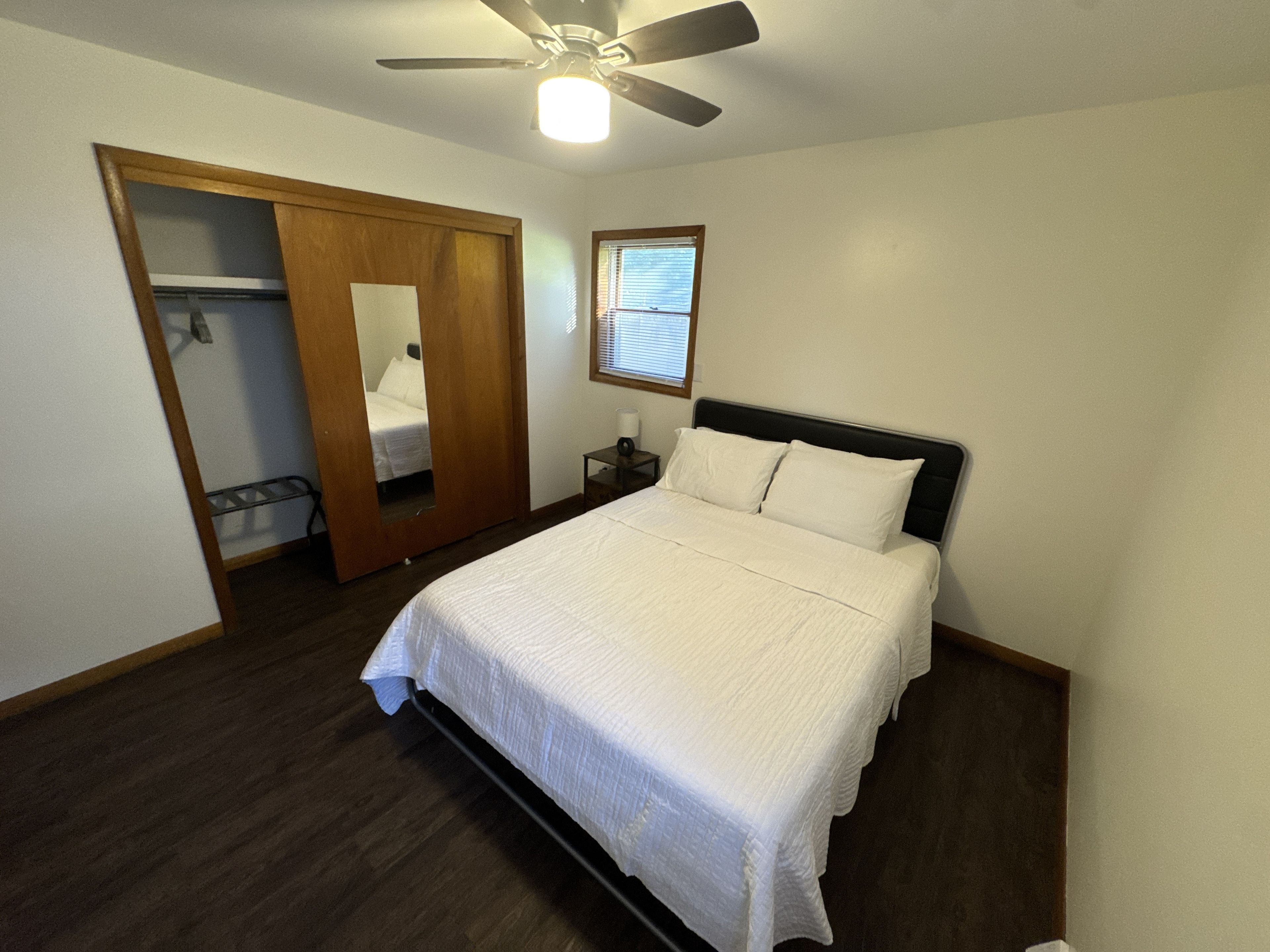 Une chambre à coucher comprenant un lit, une table de chevet, un ventilateur de plafond, une fenêtre et une porte donnant sur une autre pièce.