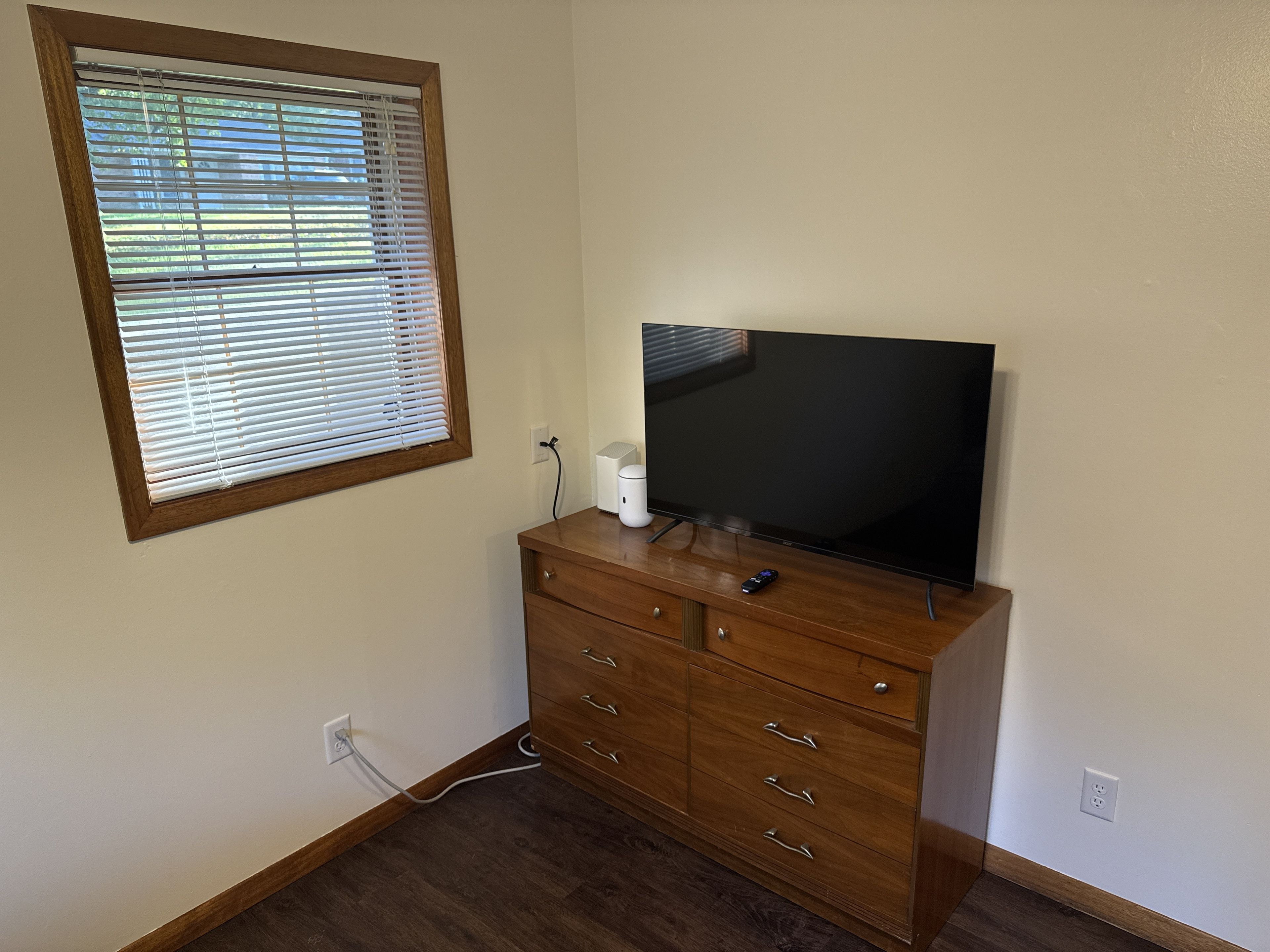 Una habitación con un aparador de madera, un televisor encima, una ventana con persianas y un enchufe en la pared.