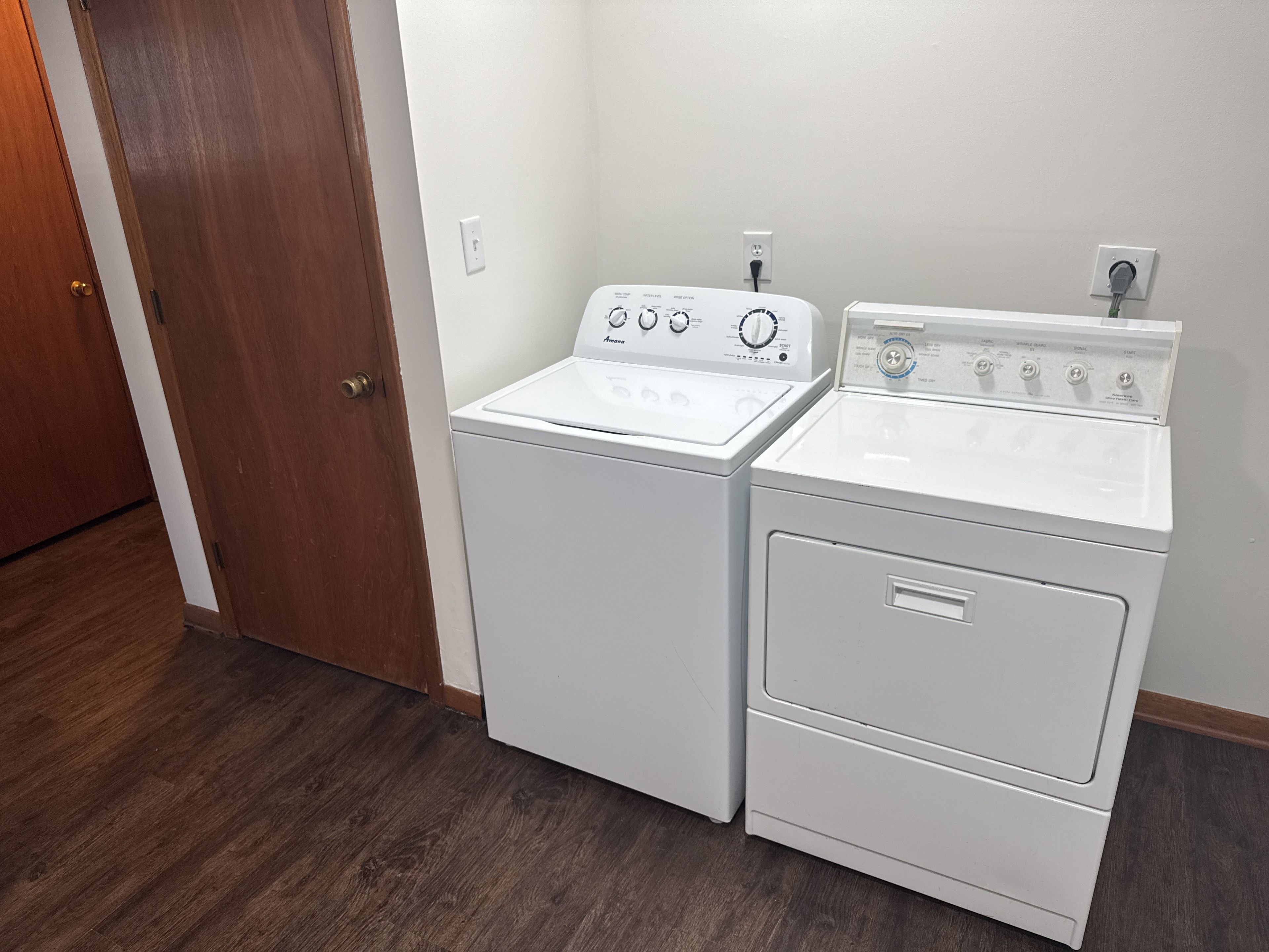 Un cuarto de lavandería con dos lavadoras y secadoras apiladas, piso de madera y una puerta.