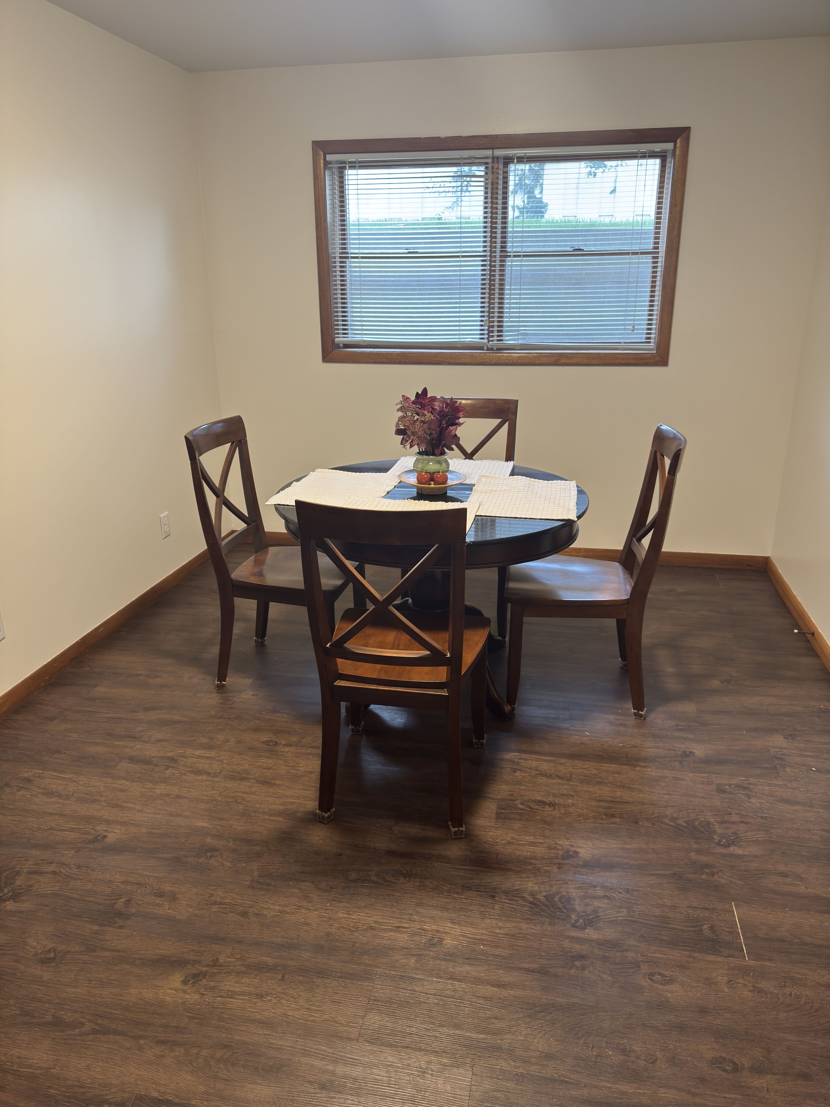 A dining area with a round table, four chairs, a vase with flowers, and a window with blinds.