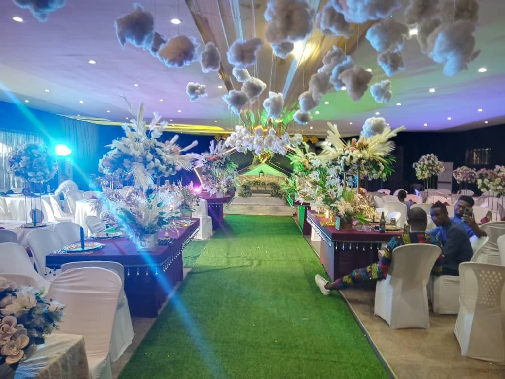 An indoor event space with a green carpet aisle, white chairs, and floral centerpieces.