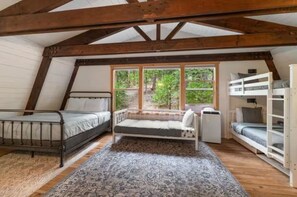 Quarto em dormitório com duas camas, uma cama de solteiro e beliche, uma janela com vista para o verde, e um tapete estampado.
