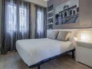 A bedroom with a bed, curtains, a bedside table, a lamp, and a wall-mounted picture. - Elegant Apartment Near Bernabeu (Madrid)