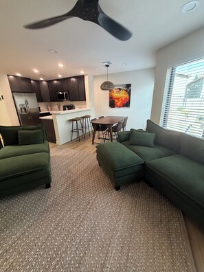 Ein modernes Wohnzimmer mit einem Ecksofa, einem Essbereich mit Tisch und Stühlen, einer Küche mit dunklen Schränken und einem Kühlschrank sowie einem Deckenventilator.